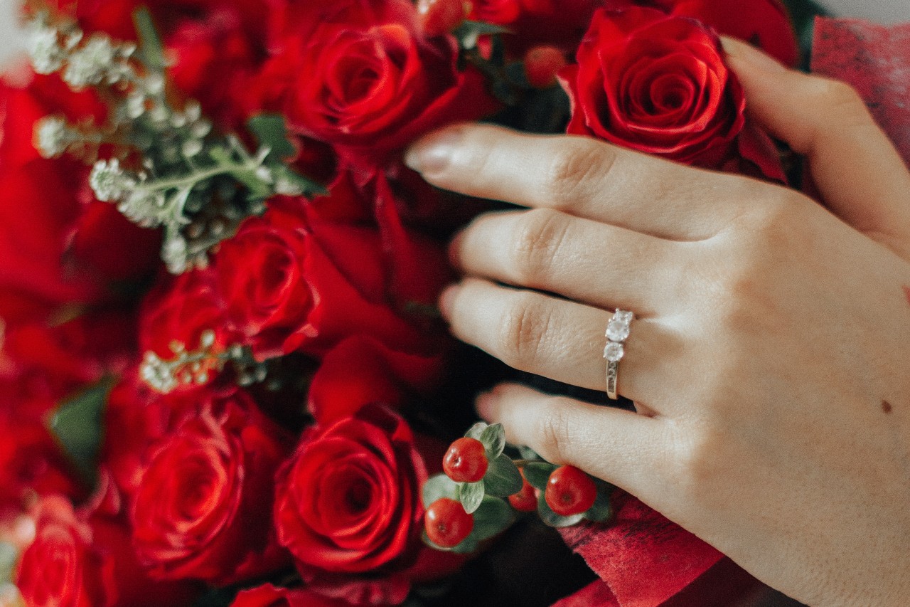 Bouquet of red roses with a woman wearing a three stone engagement ring resting her hand on the flowers
