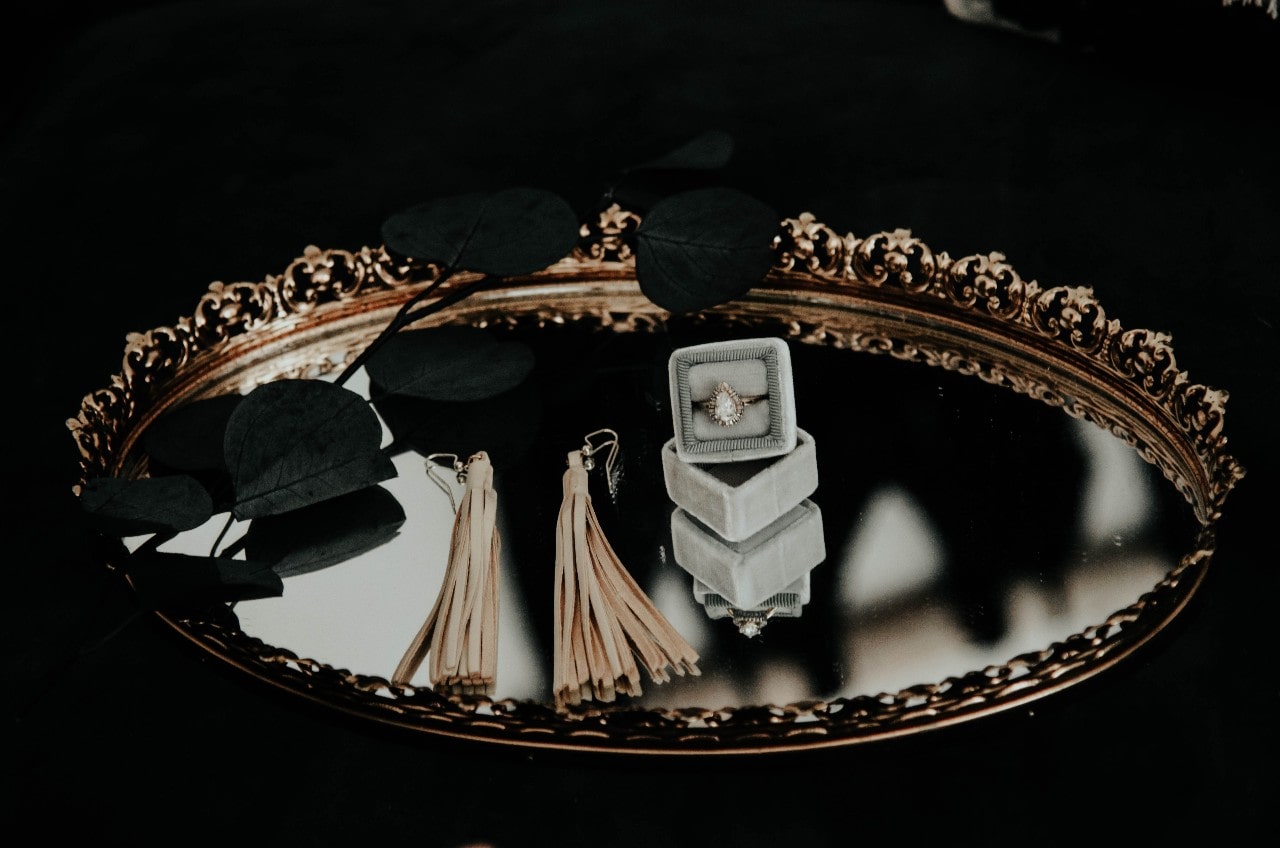 A mirror tray with a pair of earrings and a pear shape diamond engagement ring in a ring box sitting on top