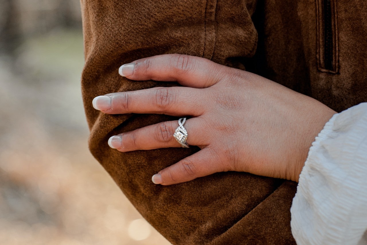 A woman’s hand resting on a jacketed arm, wearing a princess cut diamond engagement ring