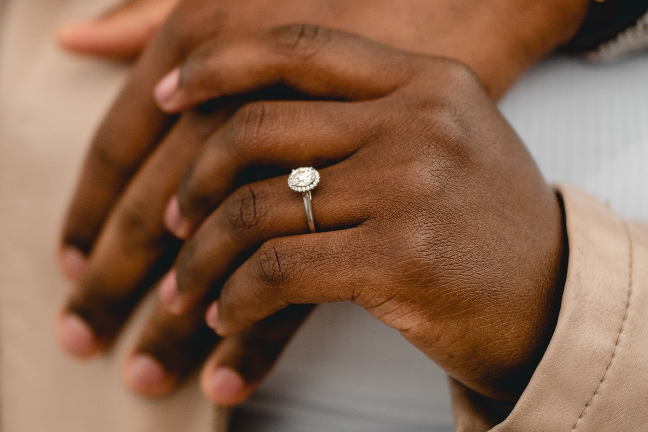 Two hands holding, one wearing a silver, oval cut halo engagement ring