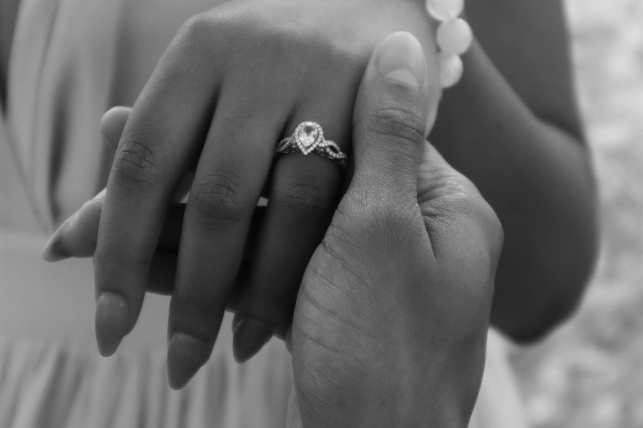 Black and white photo of a groom holding a brides hand, who is wearing a pear shape, halo engagement ring