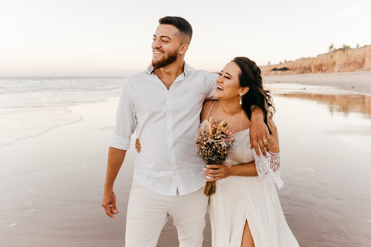 A groom and bride walk down a beach together, laughing.