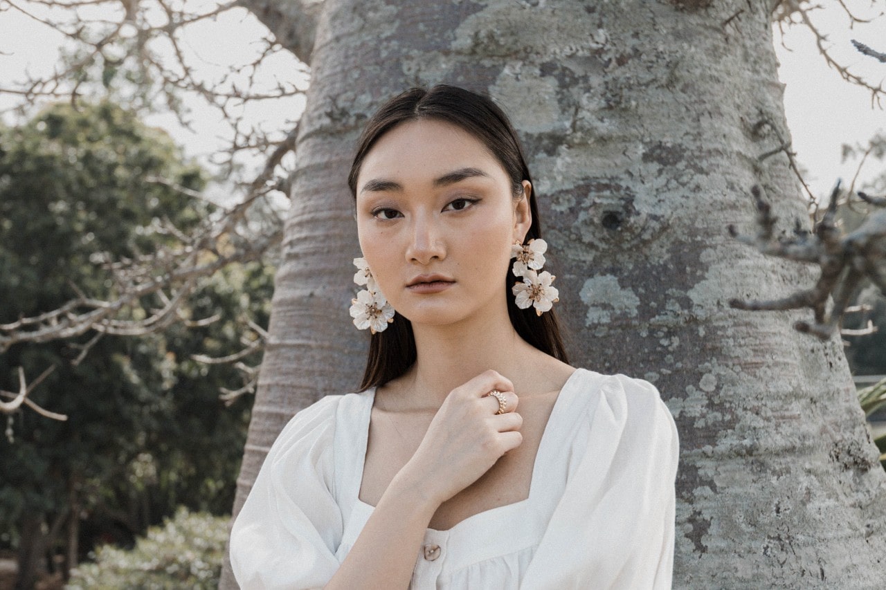 a woman standing in front of a tree, wearing a pair of white floral earrings