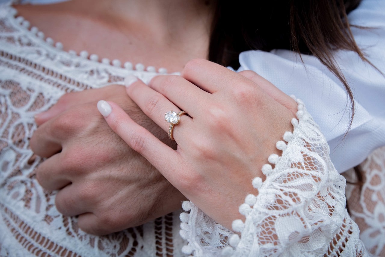 A close-up of a bride’s hand, wearing a gold engagement ring, holding her groom’s hand above her heart.