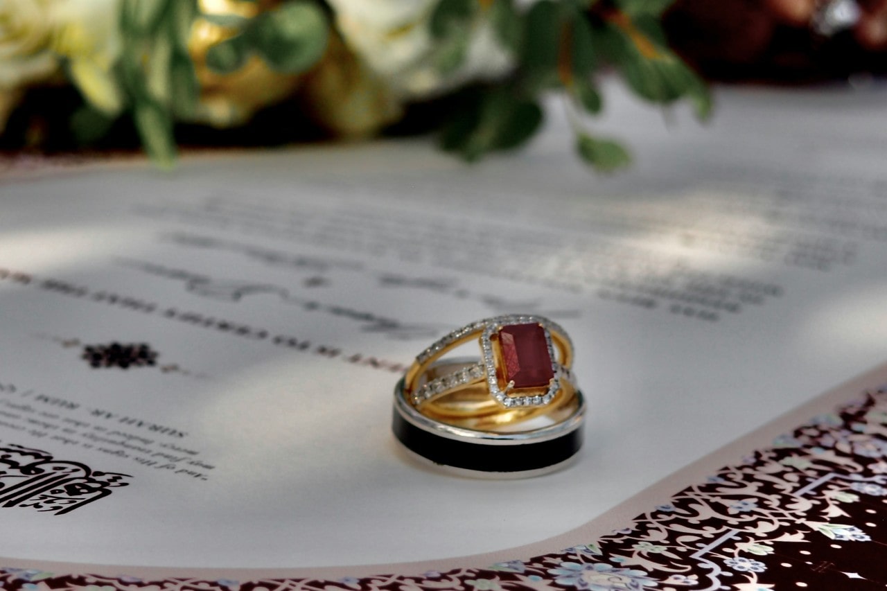 A red ruby emerald cut halo side stone engagement ring sitting inside a man’s black and silver wedding band with the matching ladies wedding band sitting on top of the engagement ring